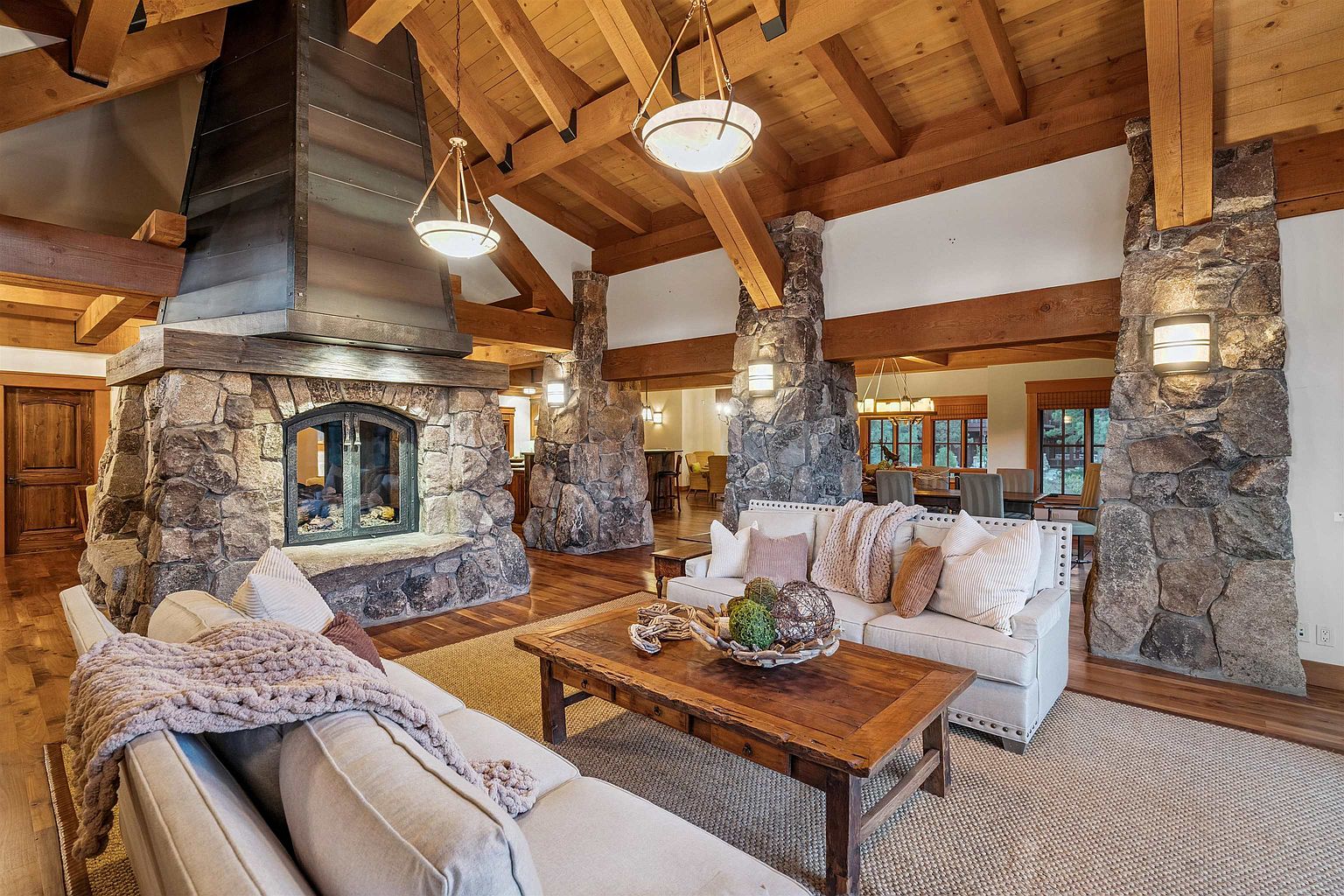 This is a spacious living room featuring a stone fireplace with a metal hood, exposed wooden beams, and stone pillars. The room is furnished with comfortable sofas and a wooden coffee table, creating a warm and inviting atmosphere. The perspective is a wide shot, showcasing the room's layout and architectural details.