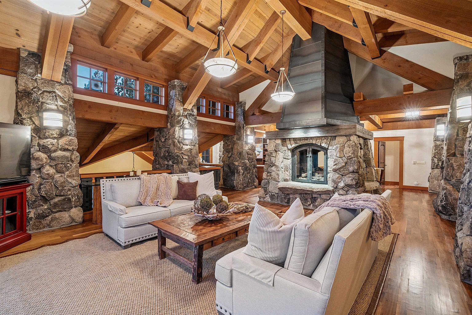 This is an interior shot of a rustic living room featuring exposed wooden beams, stone pillars, and a large stone fireplace with a metal flue. Two light-colored sofas flank a wooden coffee table, and a neutral-toned rug covers the floor. The room exudes a cozy and luxurious atmosphere, perfect for a mountain retreat.