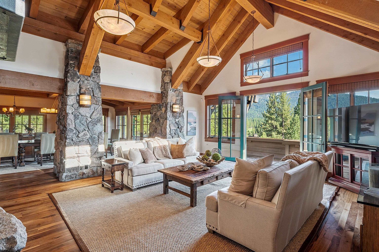 This is an interior shot of a luxurious living room featuring a high vaulted ceiling with exposed wooden beams and stone pillars. The room is furnished with comfortable sofas, a rustic coffee table, and a large area rug. Natural light floods the space through large windows and doors, offering a view of the surrounding landscape.