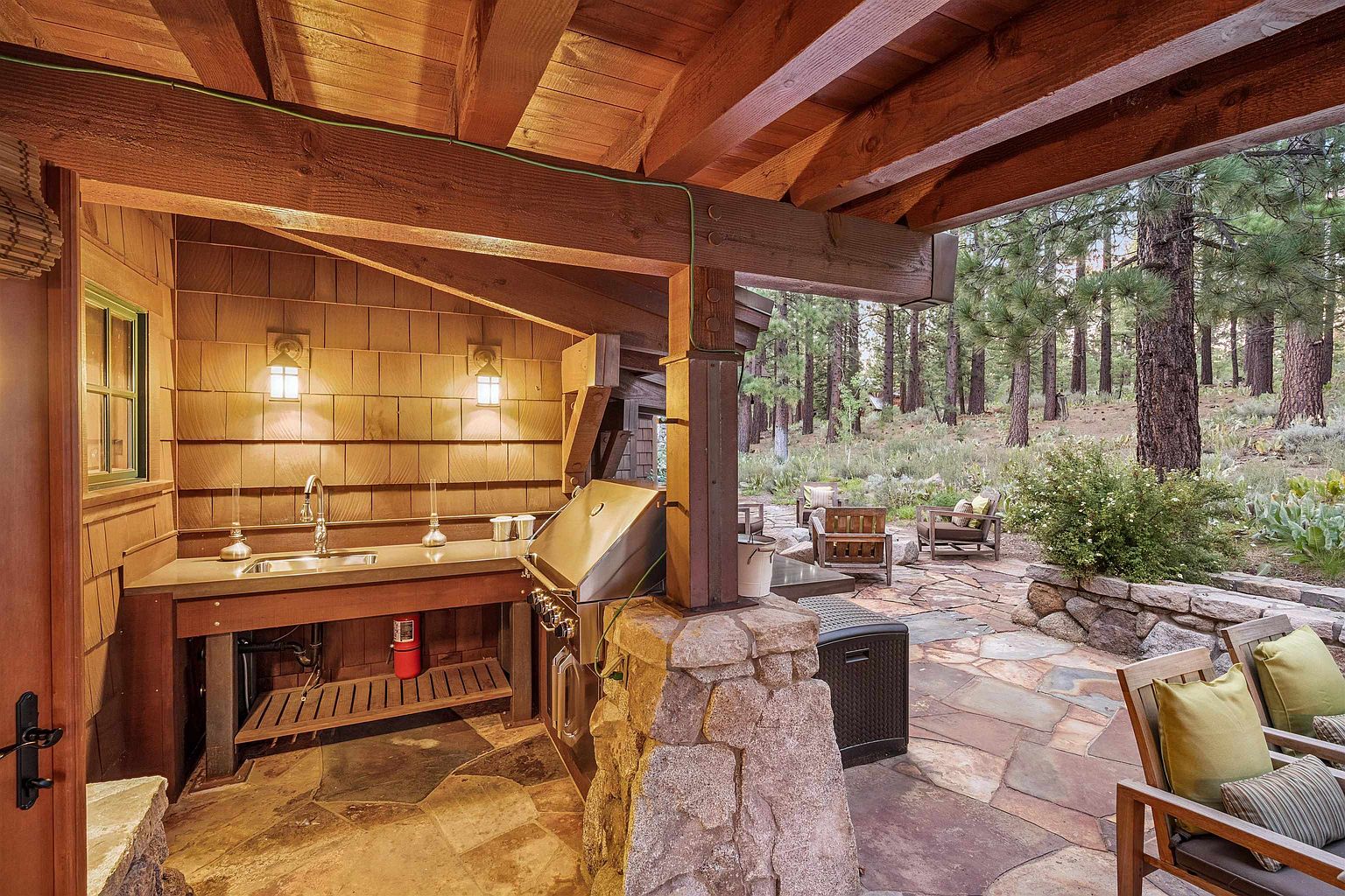 This image showcases an outdoor kitchen and patio area, perfect for entertaining. The kitchen features a built-in grill, sink, and counter space, all constructed with natural materials like stone and wood. The patio is paved with flagstone and includes comfortable seating, creating a relaxing outdoor living space with a view of the surrounding trees.