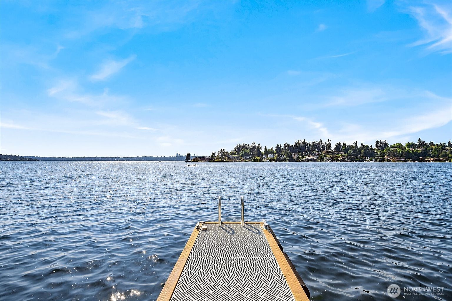 This image captures a serene, eye-level perspective looking out from a private dock onto a vast, calm lake. The dock features a textured, metallic-look surface with a sturdy metal ladder for water access, leading the viewer's eye toward the distant shoreline lined with lush trees and residential homes. The bright, expansive blue sky reflects off the rippling water, creating a peaceful and inviting atmosphere perfect for waterfront living.