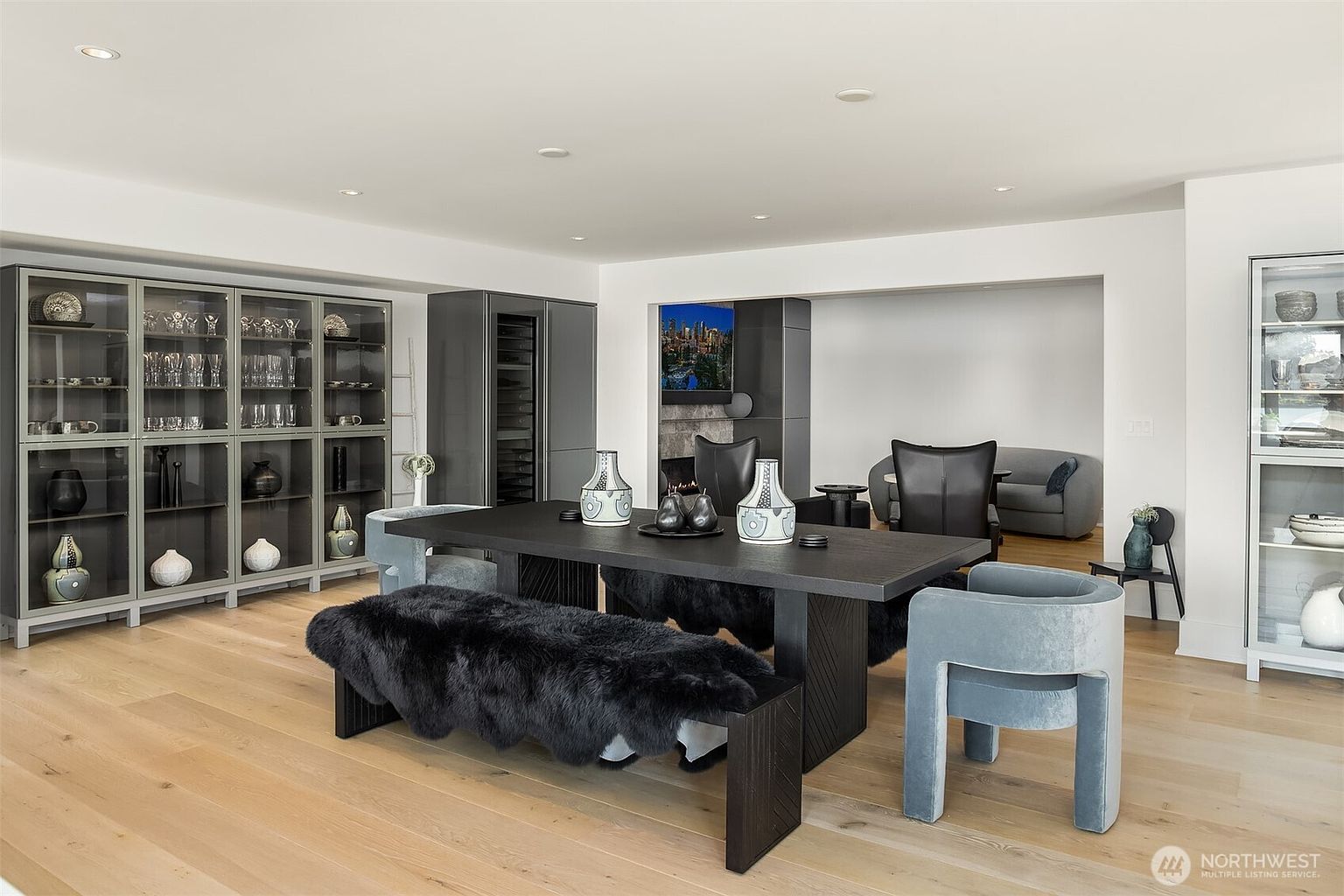 This sophisticated dining area features a large, dark-toned table paired with a plush black faux-fur bench and modern velvet-upholstered chairs. The room is anchored by extensive dark-framed glass cabinetry for display and storage, set against light wood flooring and crisp white walls. A secondary seating area is visible in the background, creating an open-concept flow that feels both luxurious and contemporary.