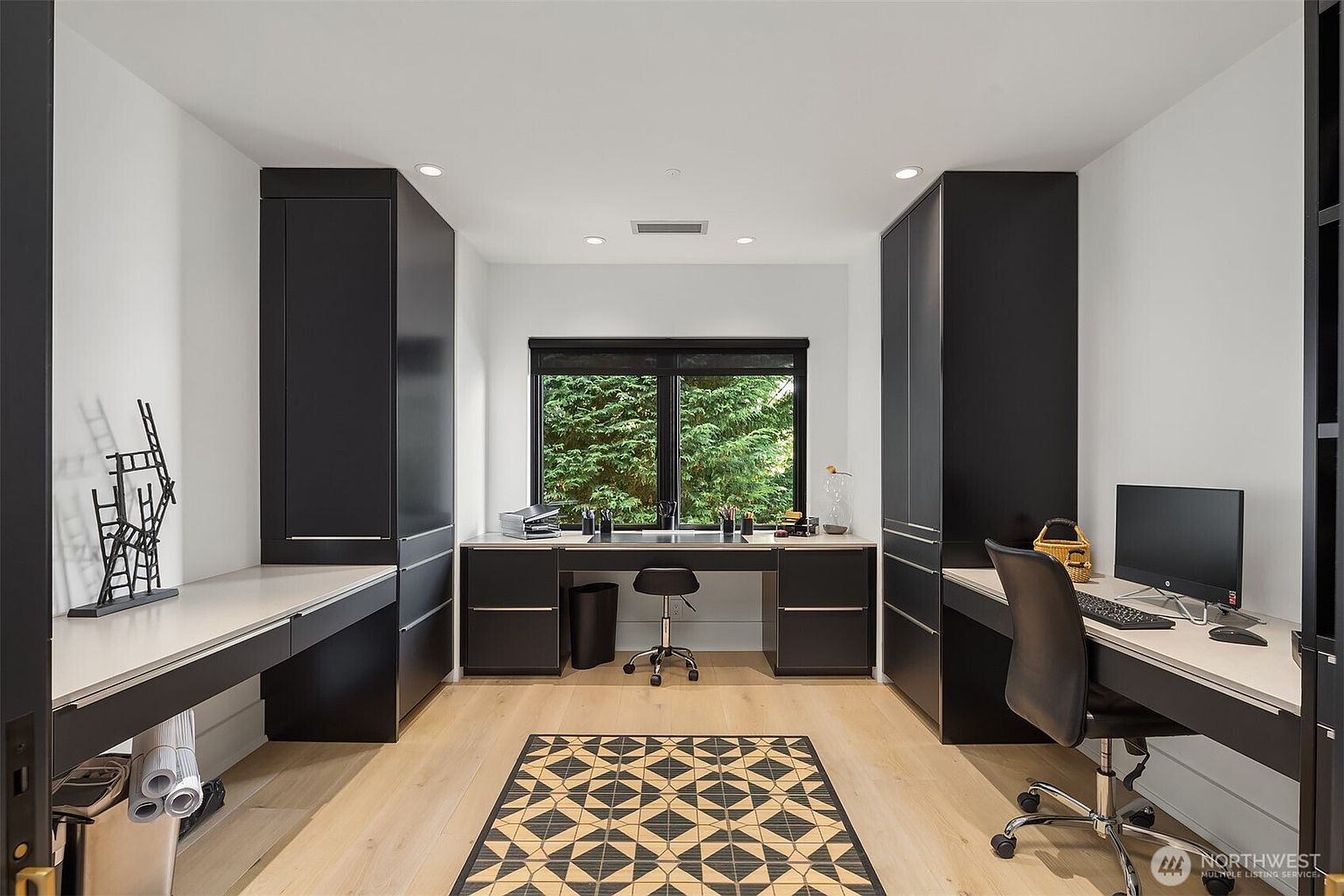 This modern home office features a symmetrical layout with dark, built-in cabinetry flanking a central window that overlooks lush greenery. The room includes two distinct desk areas with light-toned surfaces and a patterned area rug centered on the light wood flooring. The clean lines and high-contrast color palette create a professional and organized workspace.