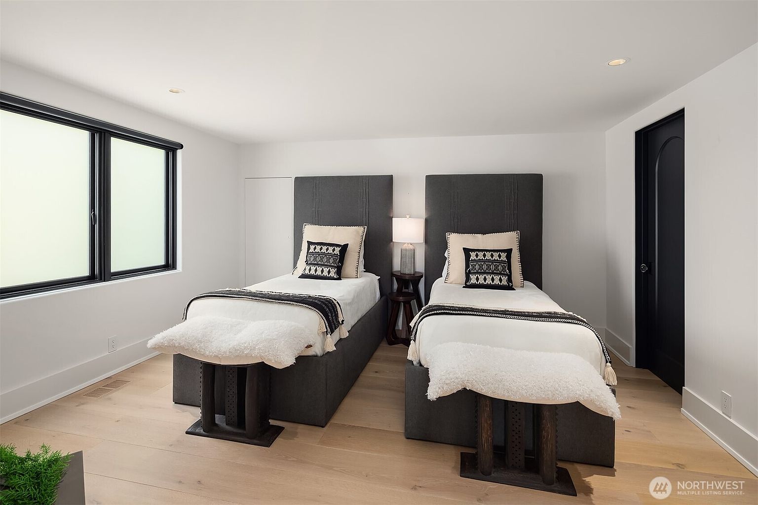 This bright and modern guest bedroom features two twin beds with tall, dark gray upholstered headboards and matching benches at the foot of each bed. The room is accented with patterned throw pillows, a central nightstand with a lamp, and light-toned wood flooring. Large windows on the left provide ample natural light, creating a clean and inviting atmosphere.