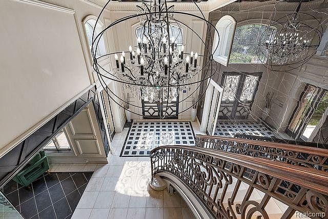 This grand foyer showcases a stunning chandelier as its centerpiece, complemented by a curved staircase with ornate metal railings. The black and white checkered flooring adds a classic touch, while the marble walls and arched windows contribute to the luxurious ambiance. The space exudes elegance and sophistication, making a memorable first impression.