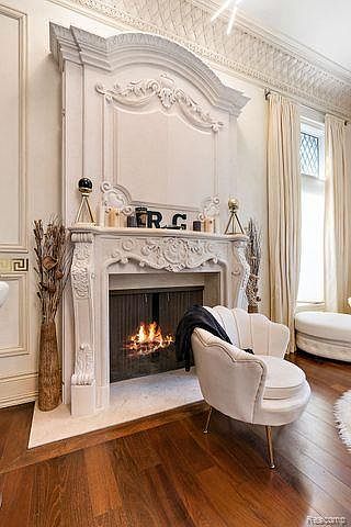 This interior shot showcases a luxurious living room centered around an ornate, white fireplace with a burning fire. A stylish, cream-colored armchair sits nearby, complementing the room's elegant aesthetic. The hardwood floors and detailed ceiling add to the room's sophisticated and inviting atmosphere.