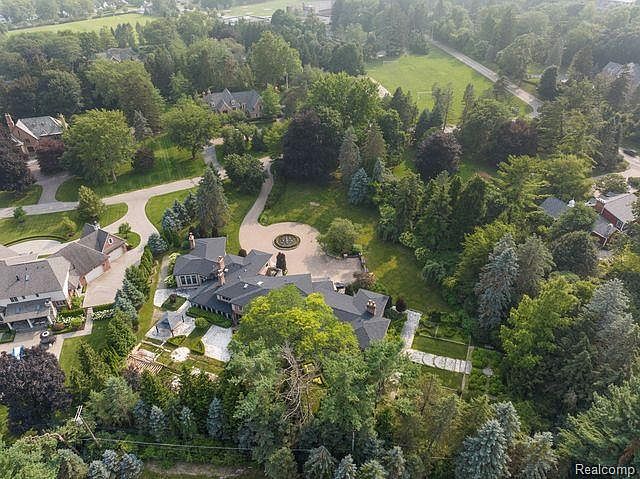This aerial view showcases a sprawling estate nestled among lush greenery. The property features a large, multi-winged house with a dark roof, surrounded by mature trees and manicured lawns. A circular driveway leads to the entrance, and a garden with geometric pathways adds to the estate's grandeur.