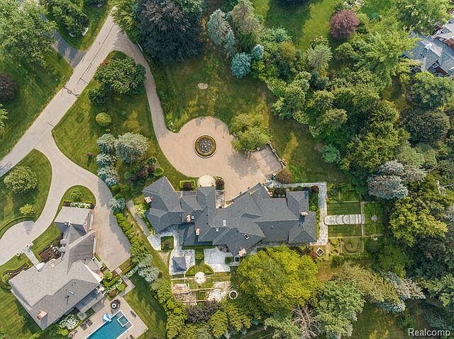 This aerial shot showcases a grand estate with a sprawling layout, featuring a large, dark-roofed house, meticulously landscaped gardens, and a circular driveway with a central fountain. The property is surrounded by lush greenery and mature trees, creating a sense of privacy and luxury. A swimming pool is visible near a secondary building, adding to the estate's appeal.