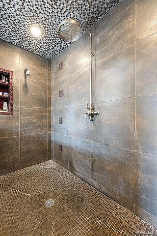 This is a primary bathroom featuring a walk-in shower with large format gray tiles and mosaic tile accents. The shower includes a rainfall showerhead and a niche for toiletries. The floor is covered in small mosaic tiles, and the overall design is modern and luxurious.