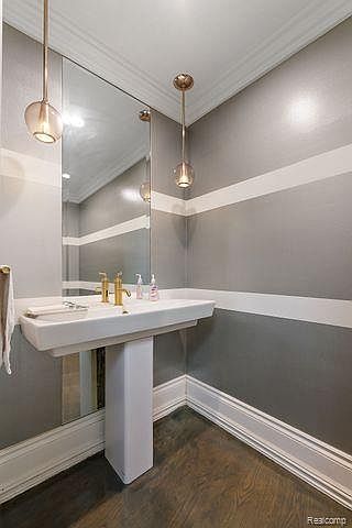 This is a well-lit powder room featuring a pedestal sink with gold fixtures, a large mirror, and stylish pendant lighting. The walls are painted in a two-tone gray with a white stripe, complemented by dark wood flooring and white trim. The space feels modern and elegant.