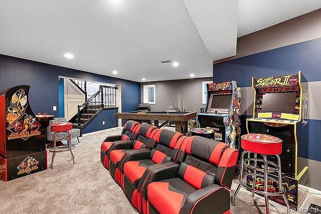 This is a basement game room featuring a pool table, arcade games, and a tiered theater seating arrangement. The walls are painted in a two-tone color scheme, and the room is well-lit with recessed lighting. The overall impression is a fun and inviting space for entertainment.