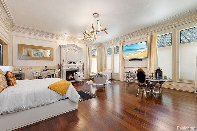 This primary bedroom features a luxurious and spacious layout with hardwood floors and ornate architectural details. A large bed is prominently placed, and a fireplace adds a cozy touch. The room also includes a seating area and a large television, creating a comfortable and elegant retreat.