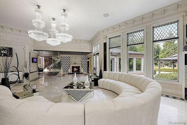 This is a luxurious living room featuring a curved white sofa, a modern geometric coffee table, and three large crystal chandeliers. The room has large windows offering views of the exterior, and the flooring is polished marble. The overall impression is one of opulence and sophistication.