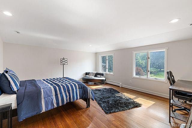 This is a spacious primary bedroom featuring hardwood floors, two large windows providing natural light, and a comfortable-looking bed with blue and white striped bedding. The room also includes a modern floor lamp, a chaise lounge, a dark-colored rug, and a desk with a chair, creating a versatile space for relaxation and work. The perspective is wide, showcasing the room's layout and size.