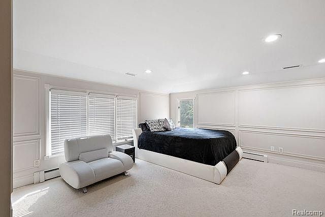 This is a spacious primary bedroom featuring a large bed with a black cover and several pillows. A modern white chair sits near the window, which is covered with blinds. The room has white walls with decorative molding and light-colored carpeting, creating a bright and airy atmosphere.