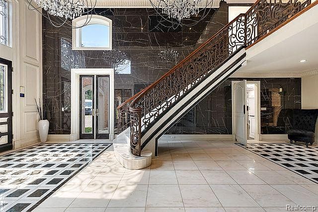 This grand foyer features a striking black and white marble floor, a sweeping staircase with ornate iron railings, and a high ceiling adorned with a crystal chandelier. A set of double doors leads to another room, and a large window provides natural light. The overall impression is one of luxury and elegance.