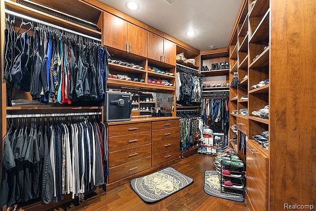 This is a well-organized walk-in closet featuring custom wood cabinetry and shelving. Clothes are neatly hung and folded, and there is a safe visible. The closet also includes shoe racks and decorative rugs, creating a luxurious and functional space.