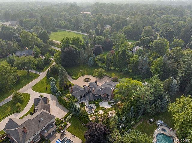This aerial view showcases a sprawling estate nestled among lush greenery. The property features a large, multi-winged house with a dark roof, a circular driveway, and meticulously landscaped gardens. A swimming pool and patio area are visible, suggesting luxurious outdoor living.