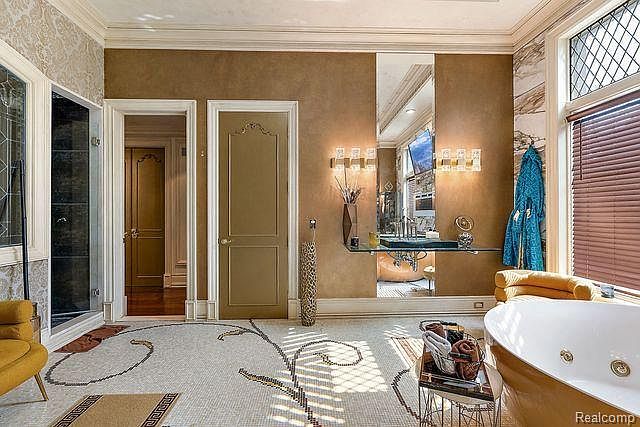 This is a luxurious primary bathroom featuring a mosaic tile floor with a floral design, a freestanding bathtub with gold accents, and a vanity area with a large mirror. The room has a sophisticated and elegant style, with warm brown walls and natural light coming through a window with blinds. The space is designed for relaxation and comfort.