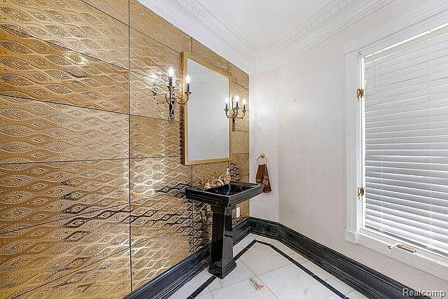 This is a stylish powder room featuring a unique gold-toned textured wall, a black pedestal sink, and a framed mirror with sconces. The floor is a combination of white and black tiles, adding to the room's sophisticated aesthetic. A window with blinds provides natural light, and the overall impression is one of luxury and elegance.