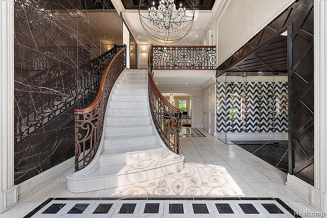 This grand foyer features a sweeping staircase with ornate iron railings and marble steps, illuminated by a dazzling chandelier. The walls are adorned with dark marble and mirrored panels, creating a luxurious and dramatic entrance. The flooring is a combination of light marble and dark accent tiles, adding to the sophisticated ambiance.