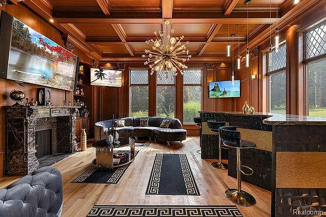 This is an interior shot of a luxurious living room featuring dark wood paneling, a coffered ceiling, and a large stone fireplace. A curved sofa anchors the space, complemented by a modern chandelier and multiple televisions. The room exudes a sophisticated and opulent atmosphere, perfect for entertaining or relaxing.