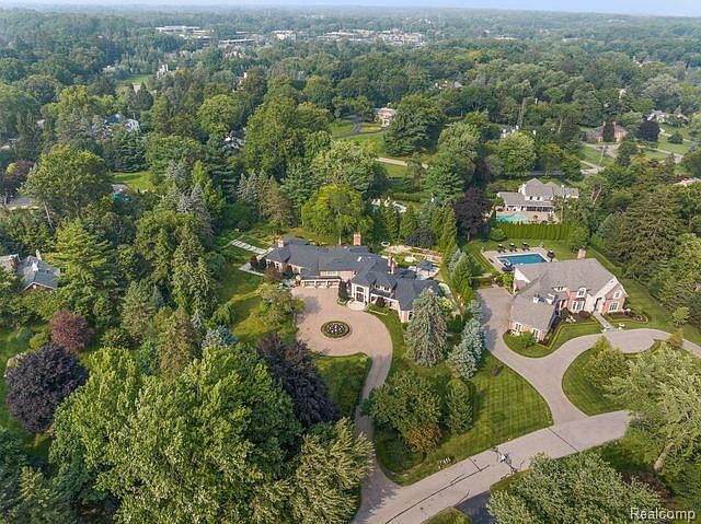 This aerial view showcases a sprawling estate nestled among lush greenery. The property features a large, well-manicured lawn, a circular driveway leading to a grand entrance, and a swimming pool. The overall impression is one of luxury and privacy, highlighting the expansive grounds and impressive architecture.