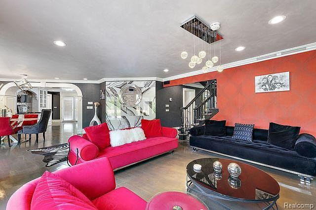 This is a vibrant living room featuring a bold color palette with a red accent wall and pink sofas. The room includes a black velvet sofa, a modern coffee table, and a unique chandelier. The space is open and stylish, with a mix of textures and patterns that create a luxurious and inviting atmosphere.