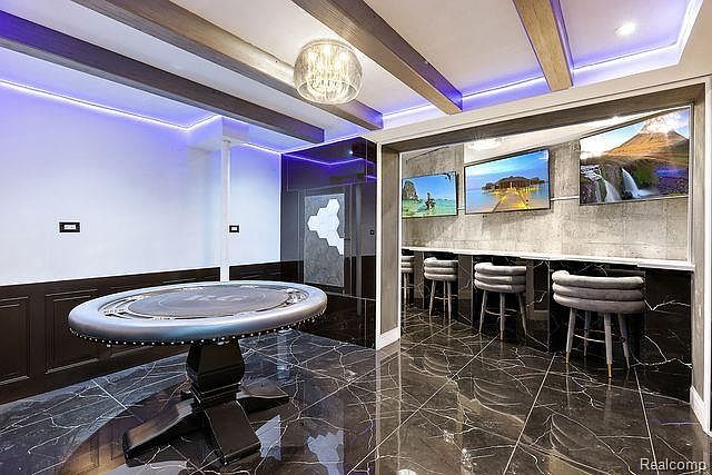 This interior shot showcases a modern living room with a unique entertainment setup. A poker table sits prominently in the foreground, while a wall-mounted display features three screens above a bar-style seating area with stools. The room is finished with dark marble flooring and accented by contemporary lighting, creating a luxurious and entertainment-focused space.