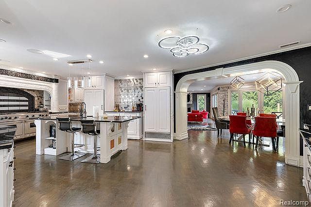 This is a spacious, open-concept kitchen and dining area featuring a large kitchen island with bar seating, white cabinetry, and modern lighting fixtures. The dining area is adjacent to the kitchen, separated by an arched entryway, and includes a dining table with red chairs. The polished floors reflect the light, enhancing the sense of space and luxury.