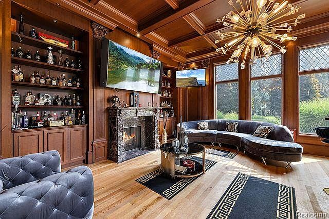 This is an interior shot of a luxurious living room featuring a large, curved sectional sofa in a plush fabric, a dark wood paneled wall with a large television above a fireplace, and a custom-built bookcase filled with decorative items and bottles. A modern chandelier hangs from the ceiling, and the room is decorated with patterned rugs, creating a sophisticated and inviting atmosphere.