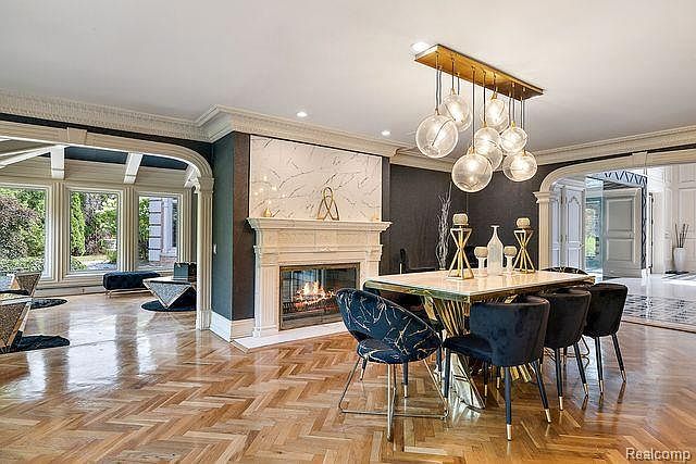 This is an interior shot of a luxurious dining room featuring a modern aesthetic. The room includes a dining table with black velvet chairs, a fireplace with a marble surround, and a unique chandelier. The herringbone wood flooring adds warmth and character to the space, creating an inviting atmosphere.