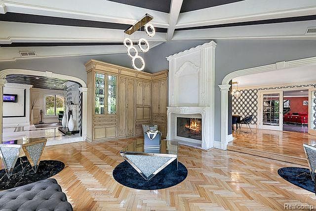 This is an interior shot of a luxurious living room featuring herringbone wood floors, a white fireplace, and custom built-in cabinetry. The room is open to other areas through arched doorways, and modern lighting fixtures add a contemporary touch. The overall impression is one of elegance and sophistication.
