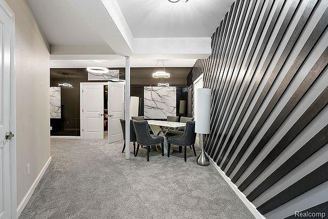 This interior shot showcases a modern dining room with a unique accent wall featuring diagonal black and white stripes. A dining table with gray chairs is centrally located, illuminated by a hanging light fixture. The room is carpeted in a neutral tone, and a glimpse into an adjacent space reveals additional architectural details and lighting.