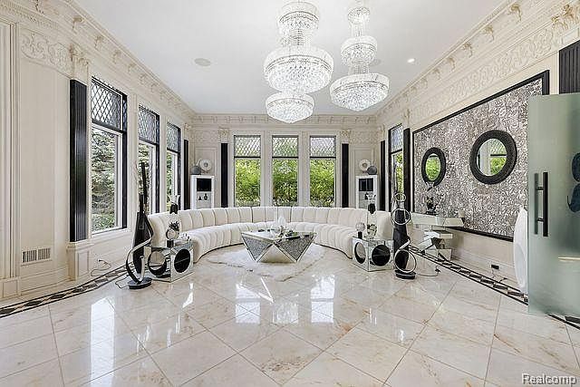 This grand living room features a curved white sofa, a geometric coffee table, and multiple crystal chandeliers, creating a luxurious and opulent atmosphere. Large windows provide ample natural light, while the marble flooring and ornate wall details add to the room's elegance. The room is designed for sophisticated entertaining and relaxation.