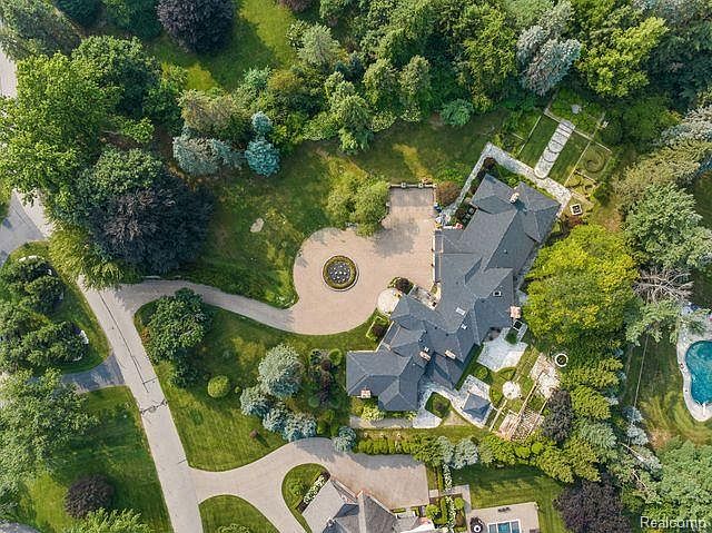This aerial shot showcases a luxurious estate with meticulously landscaped grounds, a grand circular driveway featuring a fountain, and a sprawling residence with a dark roof. The property includes a swimming pool and various garden features, all surrounded by lush greenery and mature trees, creating an impression of privacy and exclusivity.