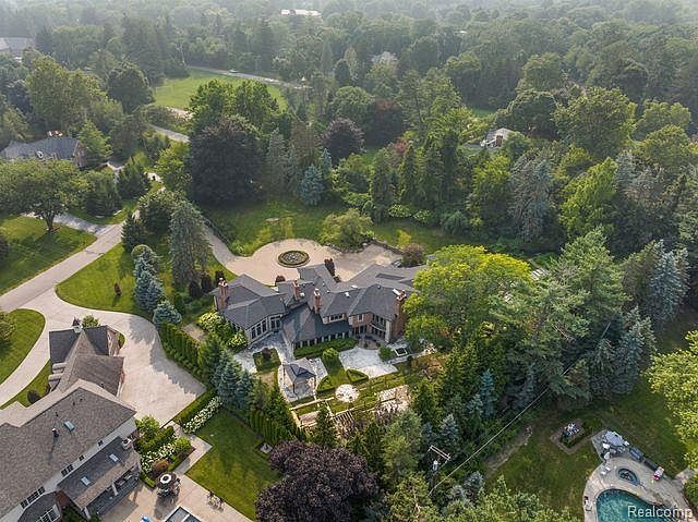 This aerial shot showcases a sprawling estate nestled among lush greenery. The large, multi-winged house features a dark roof and manicured gardens, including a formal garden area and a circular driveway with a central feature. A swimming pool and patio area are visible, suggesting luxurious outdoor living.