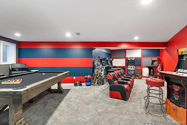 This is a vibrant basement entertainment area featuring a pool table, arcade games, and a tiered seating arrangement. The walls are painted with bold red and blue stripes, creating a playful and energetic atmosphere. The room is well-lit and carpeted, offering a comfortable space for recreation and relaxation.
