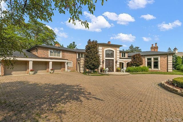 This is a front exterior view of a large, luxurious brick home. The house features a circular driveway paved with bricks, a well-manicured lawn, and mature trees. The architecture includes a prominent arched entryway and multiple garage doors, suggesting ample parking and storage space.