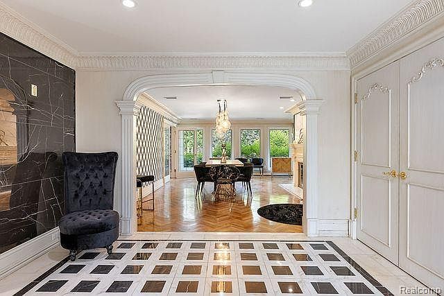 This interior shot showcases an elegant hallway leading into a dining area. The hallway features a striking black and white checkered marble floor, a high-backed black velvet chair, and a decorative black marble wall. The dining area is visible through an arched doorway, revealing a dining table with chairs and large windows that provide natural light, creating a luxurious and inviting atmosphere.