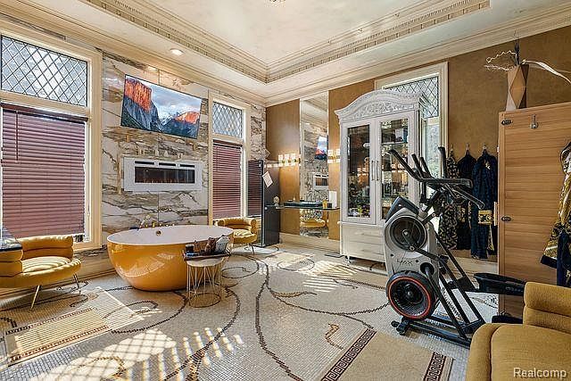 This is a luxurious primary bathroom featuring a freestanding yellow bathtub as a focal point, complemented by a modern fireplace and a wall-mounted television. The room also includes exercise equipment, suggesting a combined relaxation and fitness space. The flooring is a detailed mosaic tile, adding to the room's opulent feel.