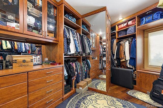 This is an interior shot of a luxurious walk-in closet featuring custom wood cabinetry and ample storage space. The closet includes hanging rods for clothing, drawers, and shelving, with a full-length mirror reflecting the organized space. A decorative rug adds a touch of elegance to the room.