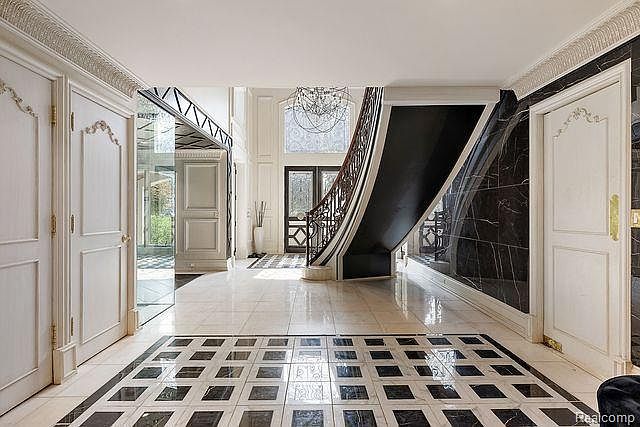 This grand foyer features a striking black and white marble tiled floor, leading to a curved staircase with dark wood accents. The walls are adorned with ornate white trim and a black marble accent wall, creating a luxurious and elegant atmosphere. A large doorway leads to an exterior view, enhancing the sense of space and light.