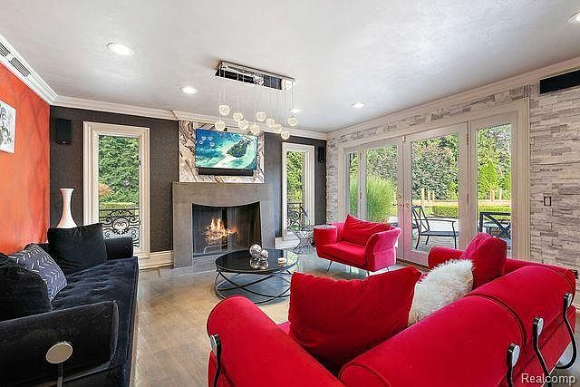 This is an interior shot of a living room featuring a black sofa, red armchairs, and a fireplace with a mounted television. The room has a modern design with a mix of textures, including a stone accent wall and a sleek coffee table. Large glass doors provide natural light and a view of the outdoor space.