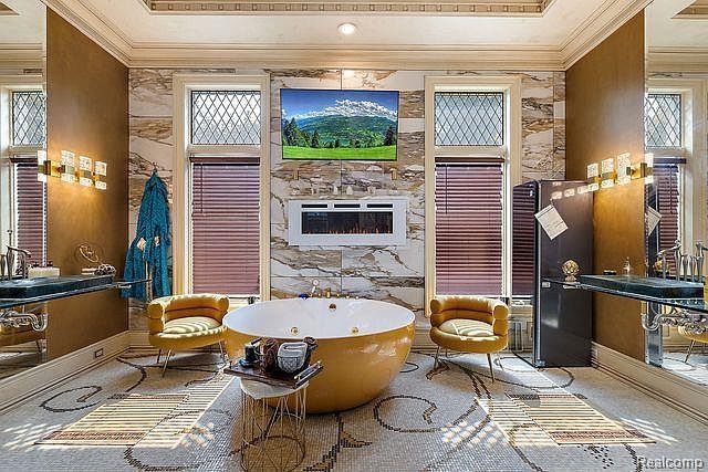 This is a luxurious primary bathroom featuring a large, round, golden bathtub as a focal point. The room is adorned with marble walls, diamond-patterned windows with blinds, and a television mounted above a modern fireplace. The mosaic tile floor adds an elegant touch, and the overall impression is one of opulence and relaxation.