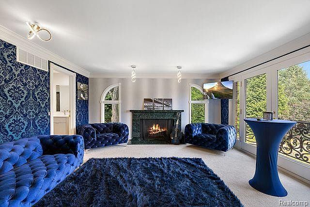 This is an interior shot of a luxurious living room featuring a blue color scheme. The room includes a tufted blue sofa, a matching area rug, and two armchairs. A fireplace is centered between arched windows, and a TV is mounted on the wall near a large window with a view of the outdoors. A blue table stands near the window.