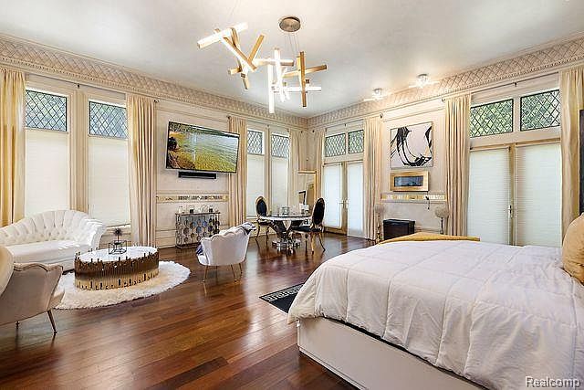 This is a luxurious primary bedroom featuring hardwood floors, ornate ceiling details, and large windows with elegant drapery. The room includes a seating area with a white sofa and a modern coffee table, as well as a dining table with chairs. A large bed dominates the foreground, suggesting a spacious and comfortable retreat.