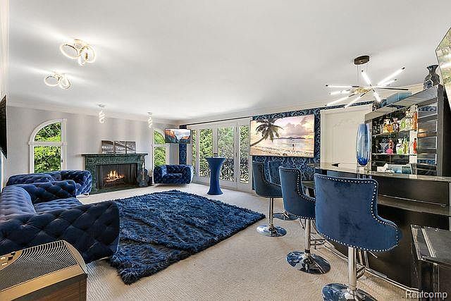 This is a spacious living room featuring a blue tufted sectional sofa, a dark blue shag rug, and a fireplace. There is a bar area with blue velvet bar stools and a large television mounted on the wall. The room has a luxurious and modern feel.