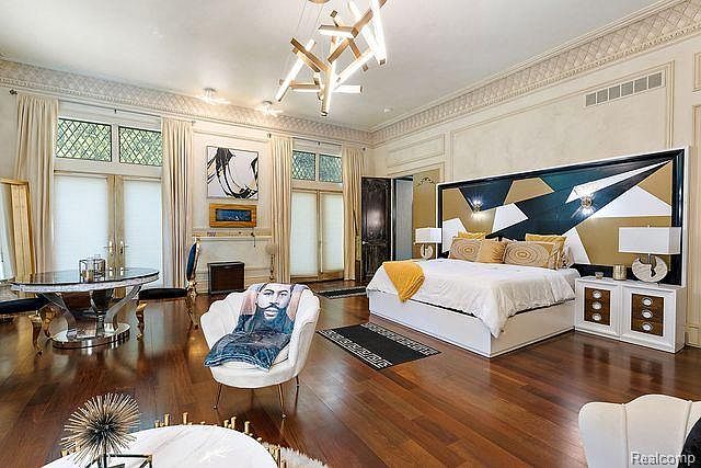 This is a spacious primary bedroom featuring hardwood floors, a unique geometric headboard, and a modern chandelier. The room includes a seating area with a round table and a decorative chair, along with large windows dressed with elegant curtains. The overall impression is luxurious and stylish.