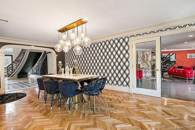 This is an interior shot of a dining room featuring a modern aesthetic. The room is furnished with a rectangular dining table, surrounded by blue velvet chairs with gold accents. A striking chandelier hangs above the table, and the walls are adorned with a geometric patterned wallpaper. The room opens to a hallway with a staircase and a glimpse into another room through a glass door.